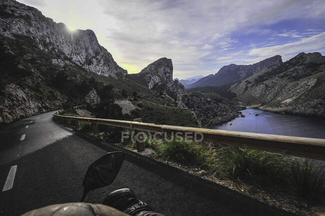 Сверху водитель урожая едет на мотоцикле по дороге, окруженной горами возле озера на фоне облачного неба — стоковое фото