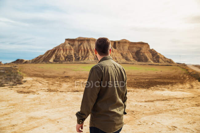 Vista posterior del hombre anónimo viajero en ropa casual de pie mirando a la gran montaña en acantilado marrón y cielo azul en el fondo en Bardenas Reales, Navarra, España - foto de stock