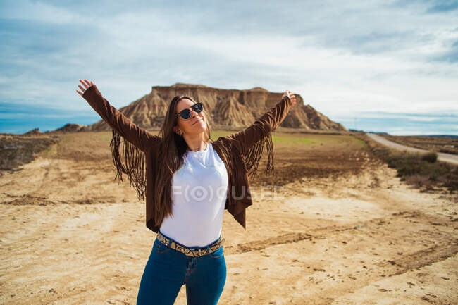 Viaggiatore donna soddisfatto in abiti casual e occhiali da sole in piedi con le mani alzate e sorridente con scogliera marrone e cielo blu sullo sfondo in Bardenas Reales, Navarra, Spagna — Foto stock