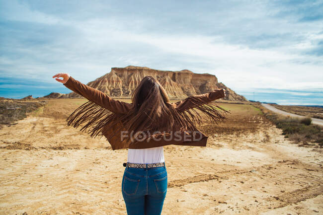 Rückansicht einer anonymen Reisenden in Freizeitkleidung, die mit erhobenen Händen in der Nähe der braunen Klippe und des blauen Himmels vor dem Hintergrund in Bardenas Reales, Navarra, Spanien steht — Stockfoto