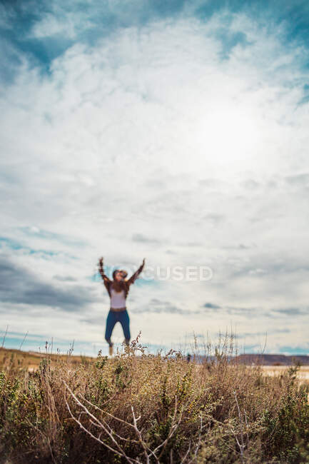 Femme méconnaissable floue en vacances dans des vêtements décontractés élégants étendant les bras dans le saut parmi la vallée déserte avec une grande falaise brune et un ciel bleu en arrière-plan à Bardenas Reales, Navarre, Espagne — Photo de stock