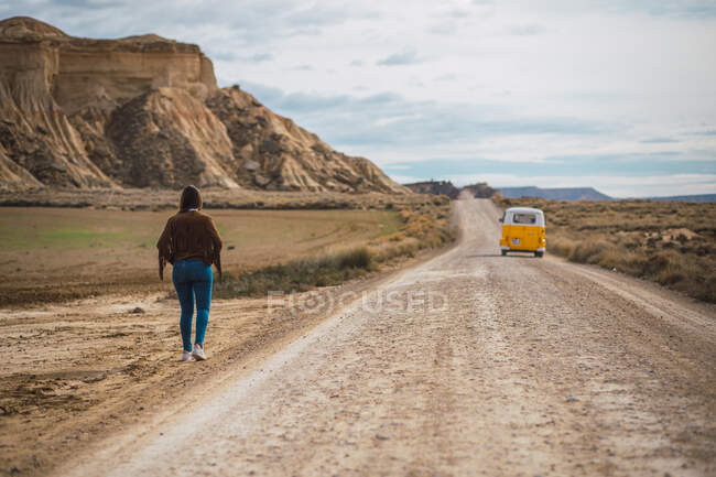 Обратный вид на неузнаваемую даму в отпуске в стильной повседневной одежде, идущую вдоль песчаной дороги среди пустынной местности и гор в Барденас-Реалес, Наварра, Испания — стоковое фото