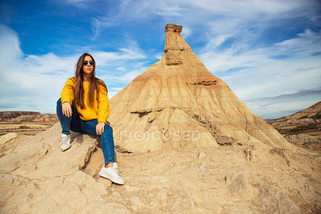 Joven viajera alegre en ropa casual elegante sentada en una colina marrón con cielo azul sobre fondo en Bardenas Reales, Navarra, España - foto de stock