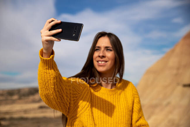Gioioso giovane viaggiatore femminile in elegante abbigliamento casual sorridente mentre prende selfie sul telefono cellulare con collina marrone e cielo blu sullo sfondo in Bardenas Reales, Navarra, Spagna — Foto stock