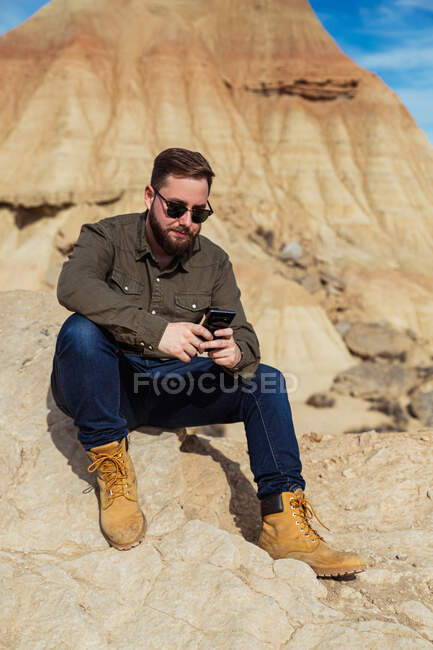 Содержание бородатый мужчина в повседневной одежде и солнцезащитные очки с помощью мобильного телефона во время отдыха у подножия коричневой горы с голубым небом на заднем плане в Bardenas Reales — стоковое фото