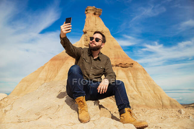 Zufriedener bärtiger Mann in Freizeitkleidung und Sonnenbrille mit Handy, während er sich am Fuße des braunen Berges mit blauem Himmel auf dem Hintergrund in Bardenas Reales ausruht — Stockfoto