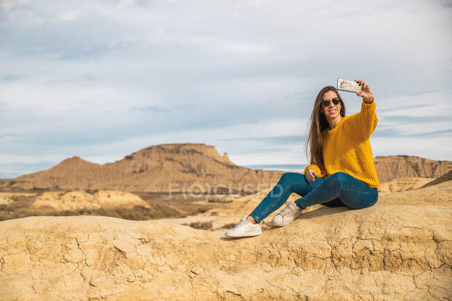 Gioioso giovane viaggiatore femminile in elegante abbigliamento casual sorridente mentre prende selfie sul telefono cellulare con collina marrone e cielo blu sullo sfondo in Bardenas Reales, Navarra, Spagna — Foto stock