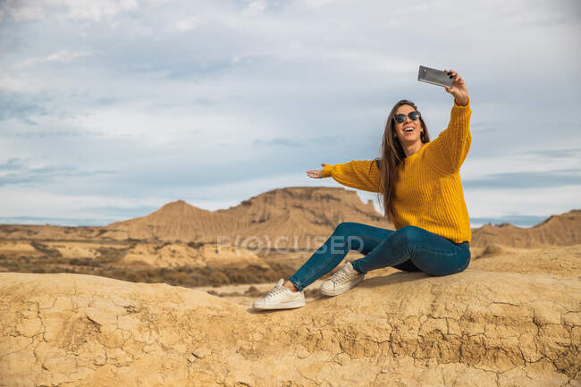 Joven viajera alegre en ropa casual elegante sonriendo mientras toma selfie en el teléfono móvil con la colina marrón y el cielo azul en el fondo en Bardenas Reales, Navarra, España - foto de stock