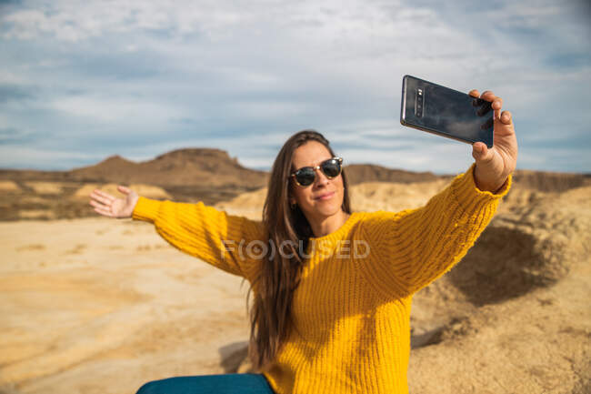 Joyeuse jeune voyageuse en tenue décontractée élégante souriante tout en prenant selfie sur téléphone portable avec colline brune et ciel bleu sur fond à Bardenas Reales, Navarre, Espagne — Photo de stock