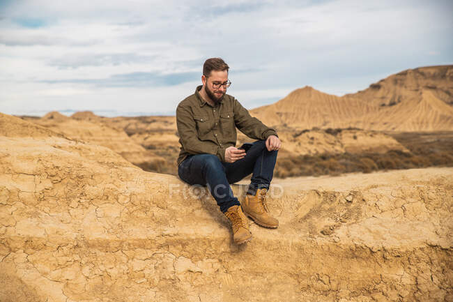 Zufriedener bärtiger Mann in Freizeitkleidung und Sonnenbrille mit Handy, während er sich am Fuße des braunen Berges mit blauem Himmel auf dem Hintergrund in Bardenas Reales ausruht — Stockfoto