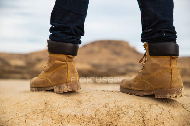 Ноги на безликому мандрівнику в коричневих чоботах і синіх джинсах стоять на брудній піщаній дорозі з горою і небом на розмитому тлі в Барденас Реалес, Наварра, Іспанія. — стокове фото