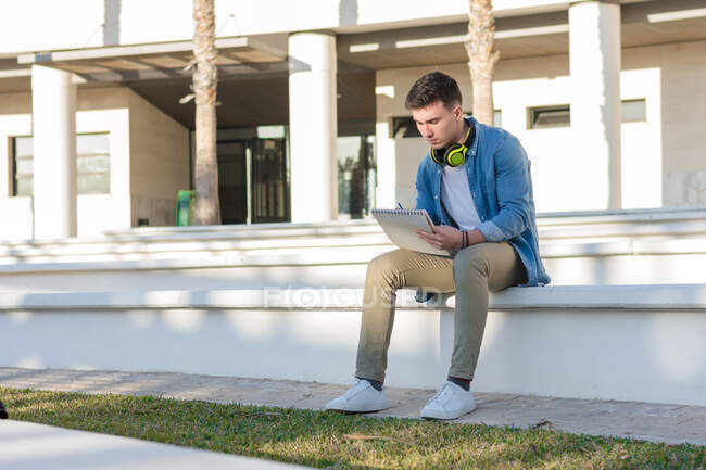 Estudiante masculino reflexivo en auriculares brillantes que estudian en la plaza de la universidad y escriben en un cuaderno sentado en la valla - foto de stock