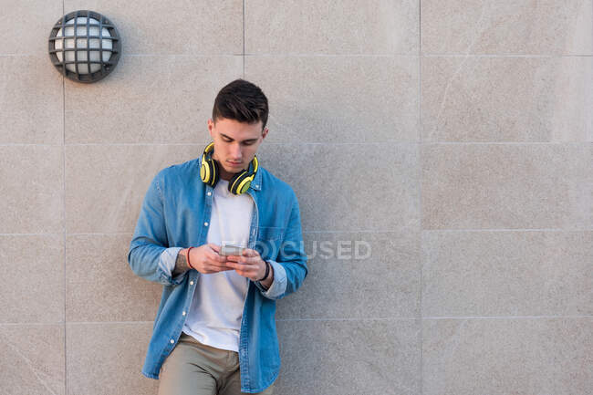 Stylish man in bright headphones surfing mobile phone while leaning on marble wall in sunny day — Stock Photo