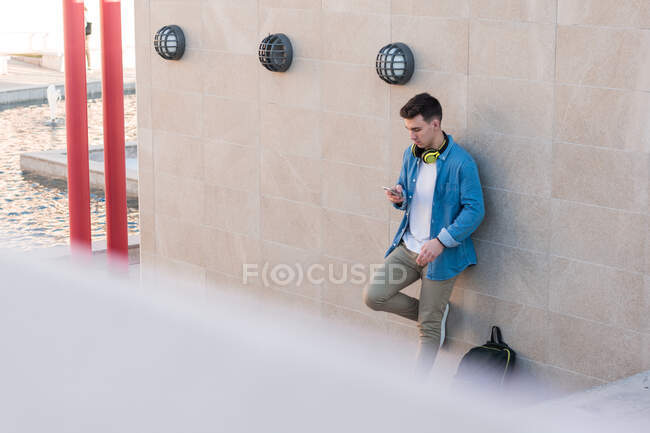 Стильний чоловік у яскравих навушниках, що серфінгують мобільний телефон, спираючись на мармурову стіну в сонячний день — стокове фото