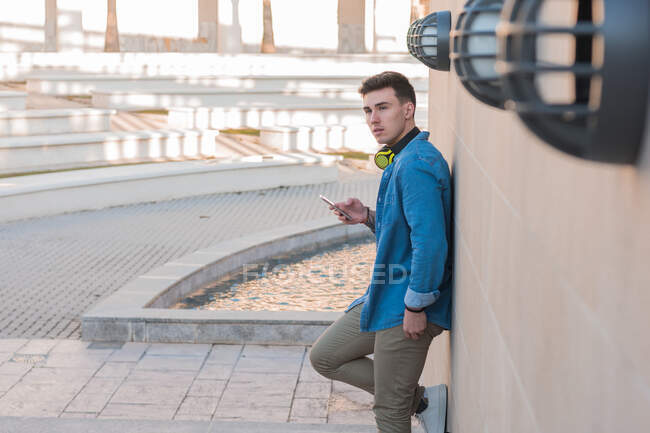 Vista lateral do homem elegante em fones de ouvido brilhantes surfando celular enquanto se inclina na parede de mármore em dia ensolarado — Fotografia de Stock