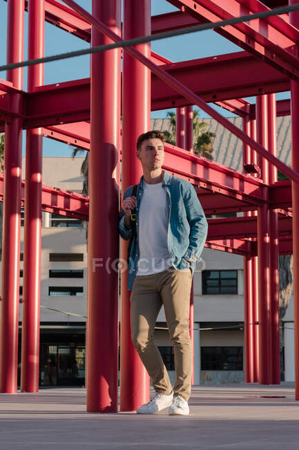 Uomo elegante con lo zaino guardando lontano in piedi in cortile con la costruzione di metallo rosso nella giornata di sole — Foto stock