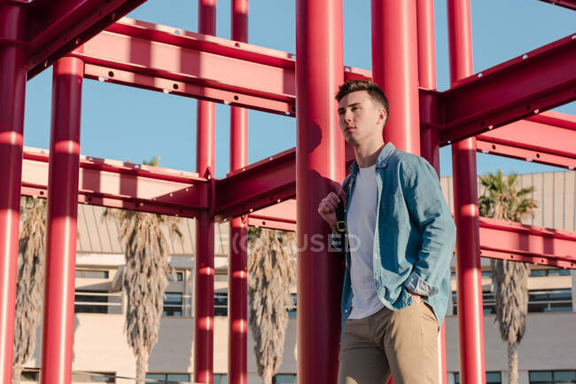Stylish man with backpack looking away standing in yard with red metal construction in sunny day — Stock Photo