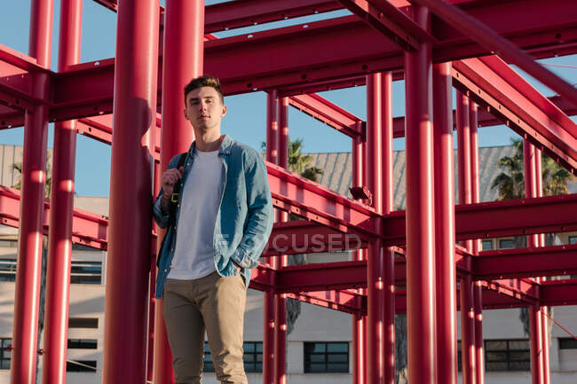 Stilvoller Mann mit Rucksack blickt in die Kamera, die im Hof mit roter Metallkonstruktion bei sonnigem Tag steht — Stockfoto