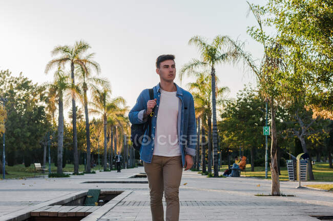 Zufriedener stilvoller Mann mit Rucksack spaziert die Parkallee entlang und schaut bei sonnigem Tag weg — Stockfoto