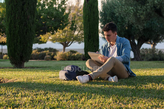 Концентрированный мужчина с рюкзаком учится писать на блокноте, сидя в парке на траве со скрещенными ногами в солнечный день — стоковое фото