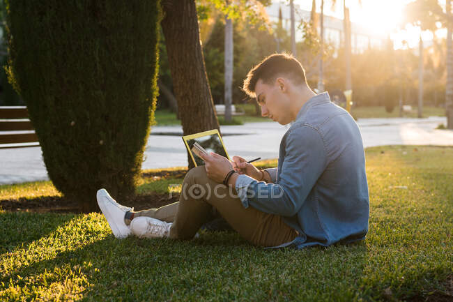 Side view of concentrated man with backpack studying at laptop and writing on a notepad sitting in park grass with crossed legs in sunny day — Stock Photo