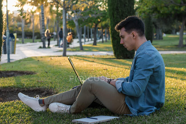 Concentrated man with backpack studying at laptop sitting in park grass with crossed legs in sunny day — Stock Photo