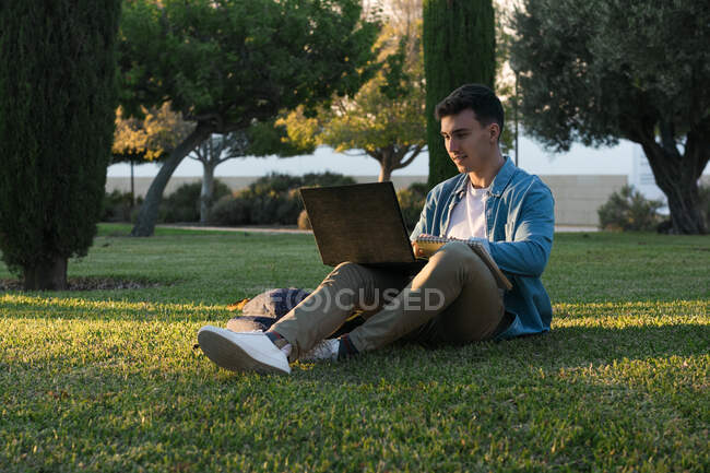 Homem concentrado com mochila estudando no laptop sentado na grama do parque com pernas cruzadas no dia ensolarado — Fotografia de Stock