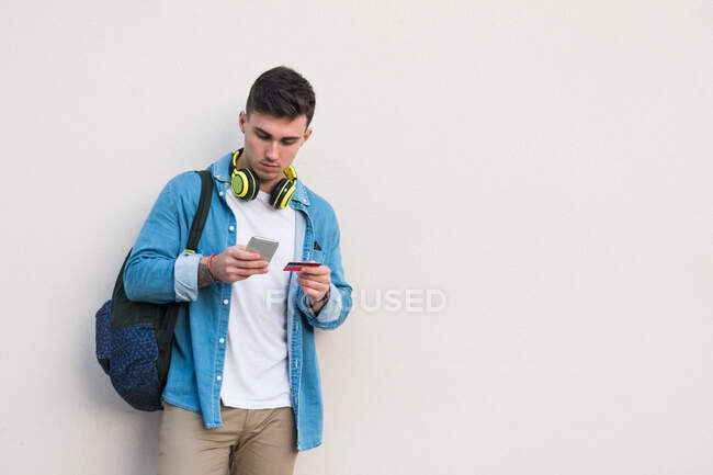 Стилістичний чоловік у яскравих навушниках серфінг мобільного телефону і тримає кредитну картку, спираючись на стіну мармуру в сонячний день. — стокове фото