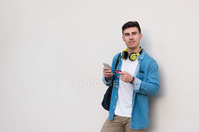 Стильний чоловік в яскравих навушниках, що серфінгують мобільний телефон і тримають кредитну картку, спираючись на мармурову стіну — стокове фото
