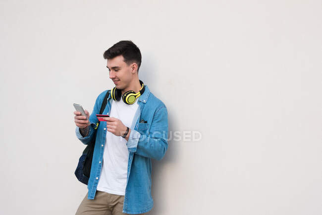 Homem elegante em fones de ouvido brilhantes surfando telefone celular e segurando um cartão de crédito enquanto se inclina na parede de mármore em dia ensolarado — Fotografia de Stock