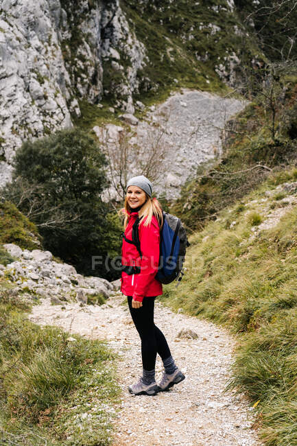 Seitenansicht einer aktiven Wanderin in roter Jacke mit schwerem Rucksack, die in die Kamera schaut, während sie im Gebirge in den Gipfeln Europas, Asturiens, Spanien wandert — Stockfoto