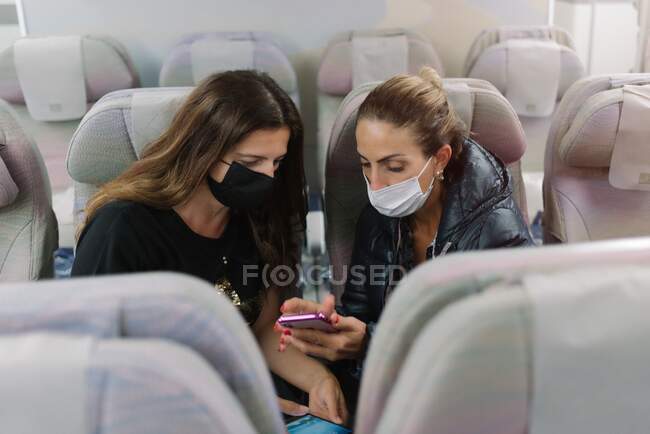 From above of female in mask showing cellphone to friend sitting in seat in airplane — Stock Photo