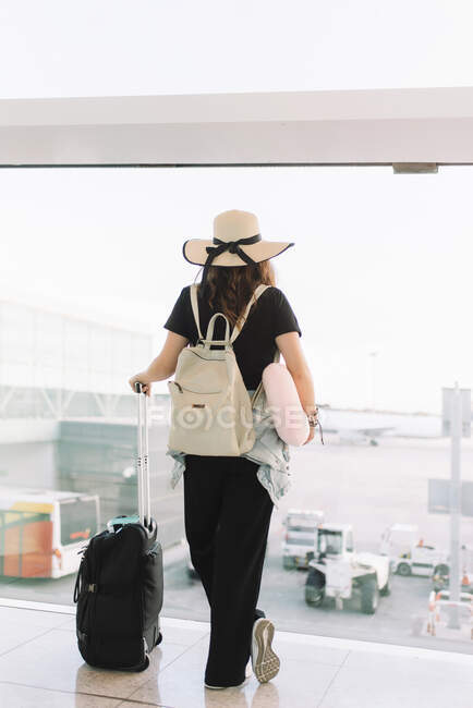 Вид ззаду леді в повсякденному одязі і капелюсі з багажем, який чекає вильоту в аеропорту, дивлячись у вікно — стокове фото