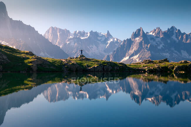 Turista solitario en la costa montañosa reflejándose en el lago de cristal en las montañas nevadas a la luz del sol - foto de stock