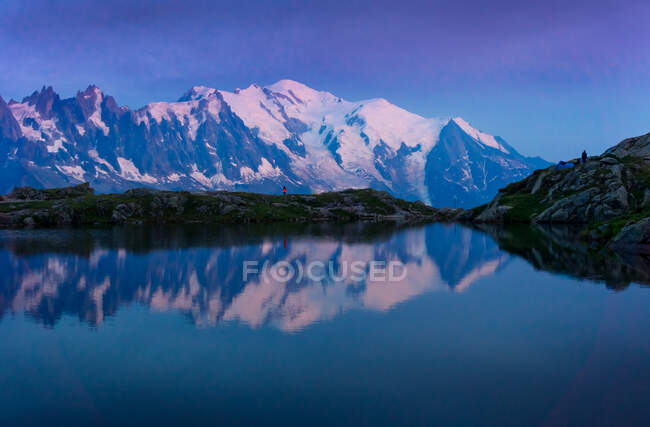 Turista solitario en la costa montañosa reflejándose en el lago de cristal en las montañas nevadas a la luz del sol - foto de stock