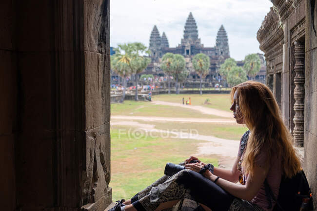 Side view of concentrated young woman with camera contemplating ancient religious temple while resting in Angkor Wat in Cambodia — Stock Photo