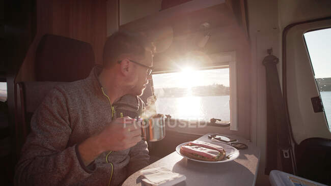 Hombre concentrado en ropa casual mirando el amanecer desde la ventana mientras está sentado y desayunando en vehículo por la mañana en Irlanda con luz trasera - foto de stock
