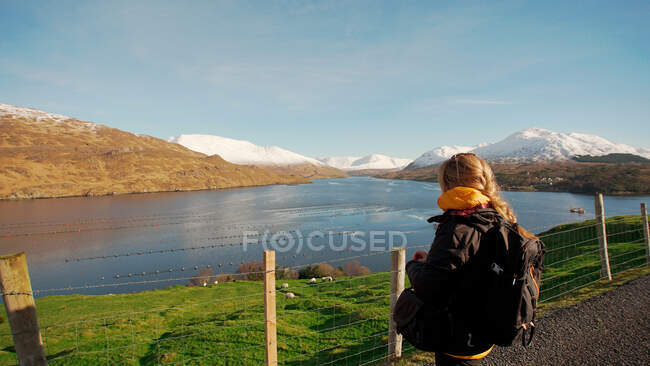 Vista posterior de la mujer sin rostro con la mochila contemplando el río y las cadenas montañosas mientras está de pie en la valla en la carretera en Irlanda - foto de stock