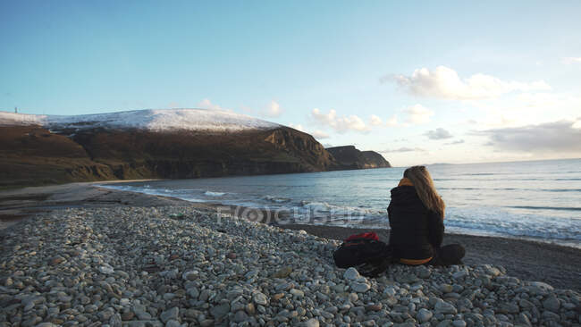 Back view of unrecognizable tourist woman contemplating nature landscape while sitting on stony beach on coastline of Ireland — Stock Photo