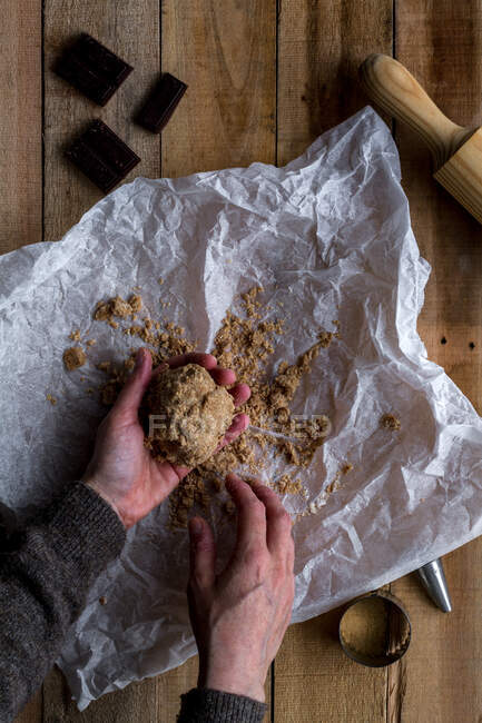 De dessus la personne de récolte tenant la pâte de chocolat à la main au-dessus des moules à biscuits en métal de chocolat de papier de cuisson blanc et rouleau sur la table en bois — Photo de stock