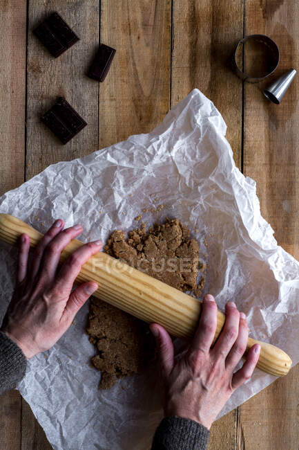 De arriba persona de la cosecha rodando masa de chocolate con rodillo sobre papel de hornear blanco chocolate con moldes de galletas de metal en la mesa de madera - foto de stock