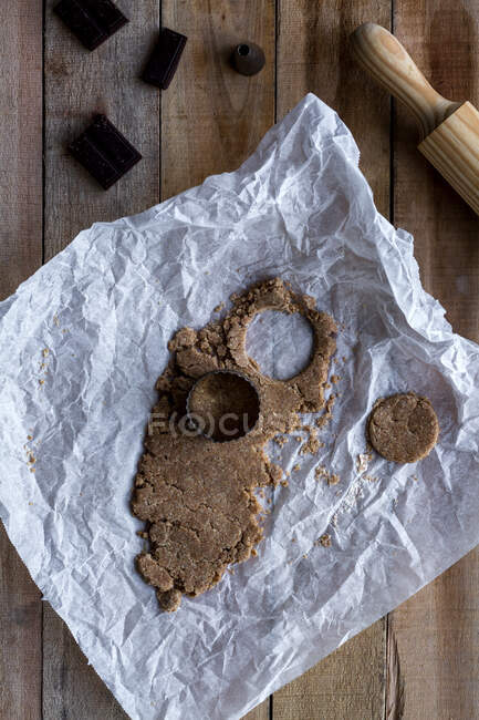 From above metal cookie mold and shape in chocolate dough on white baking paper with chocolate and rolling pin on wooden table — Stock Photo