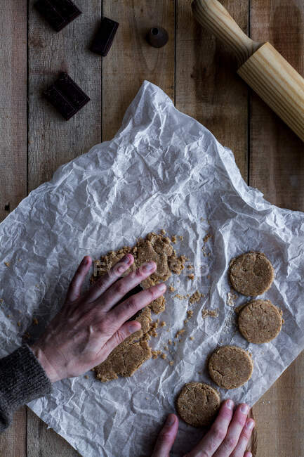 Зверху врожаю людина тримає шоколадне тісто в руці над білим папером для випічки шоколадних металевих форм печива та прокатного штифта на дерев'яному столі — стокове фото