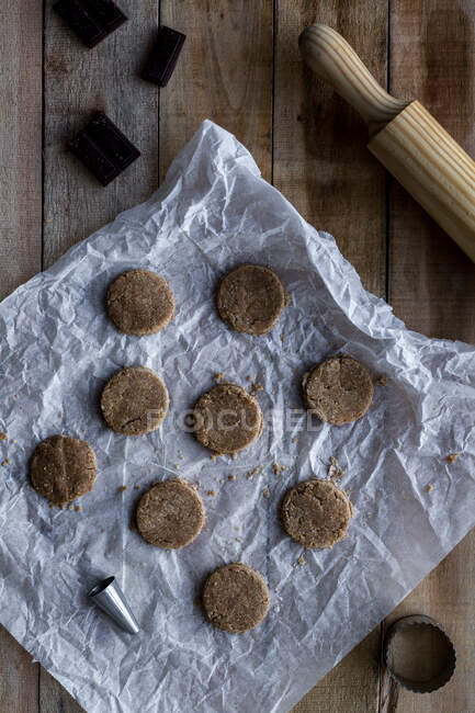 From above chocolate unbaked round cookies on white baking paper with chocolate metal cookie molds and rolling pin on wooden table — Stock Photo