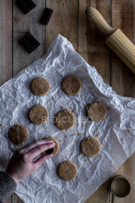 From above crop person holding chocolate dough in hand above white baking paper chocolate metal cookie molds and rolling pin on wooden table — Stock Photo