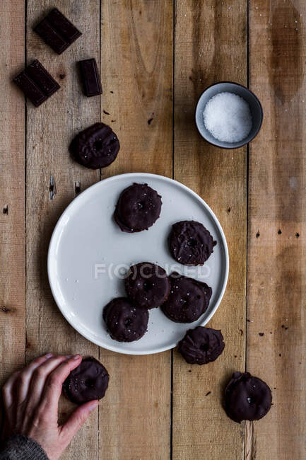 From above cropped anonymous hand grabbing aromatic cookies covered with chocolate syrup in white plate on wooden table — Stock Photo