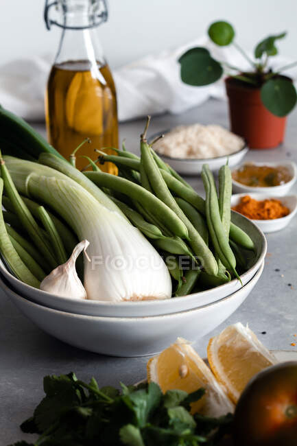 De arriba cebolla verde con ajo y judías verdes en tazón y limón con tomates y queso en plato y condimentos en la mesa - foto de stock