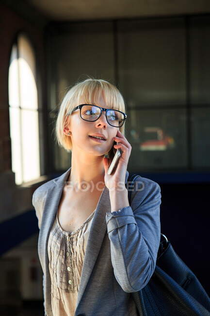 Junge blonde Geschäftsfrau mit Brille spricht auf Smartphone und schaut weg — Stockfoto