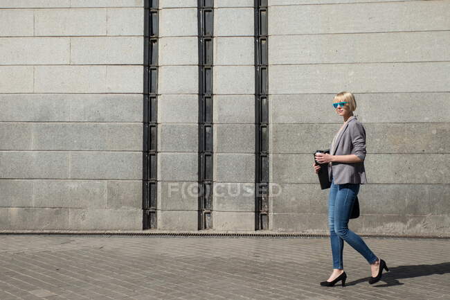 Vista lateral de empresária loira bonita em casaco formal andando com pasta e copo de papel na parede de concreto na rua ensolarada — Fotografia de Stock