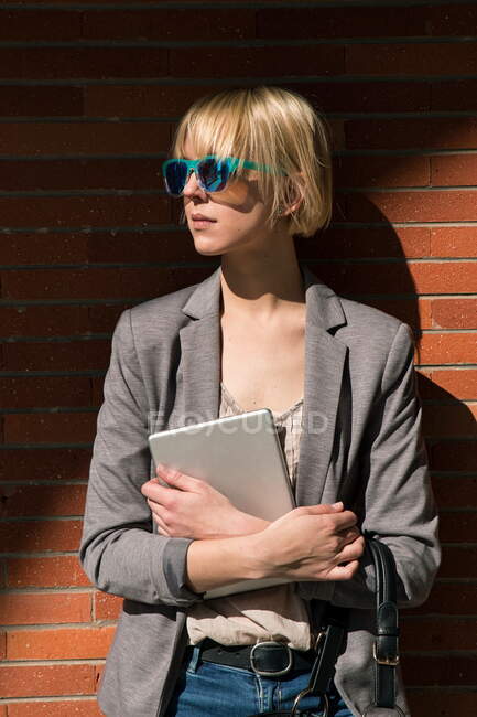 Mujer de negocios rubia en gafas de sol sosteniendo la tableta y apoyado en la pared de ladrillo - foto de stock